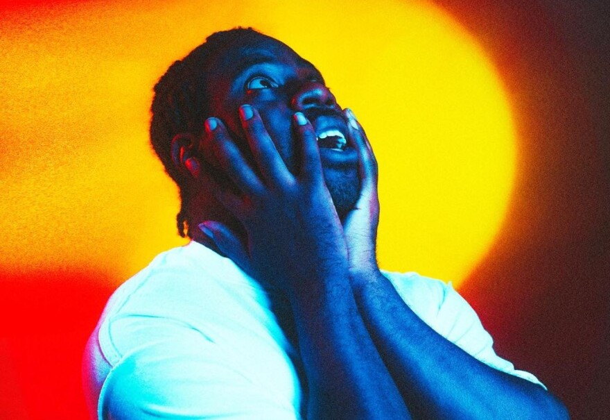 A Black man wearing a white t-shirt holds his face while standing in front of a colorful background