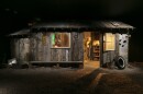 A small ramshackle cabin made out of wood sits in an exhibit in an art museum. The cabin is well-lit, but the surrounding space is made to look like night.