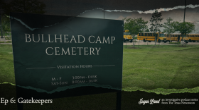 A sign in the foreground reads "Bullhead Camp Cemetery." In the background, yellow school busses fill a parking lot. 