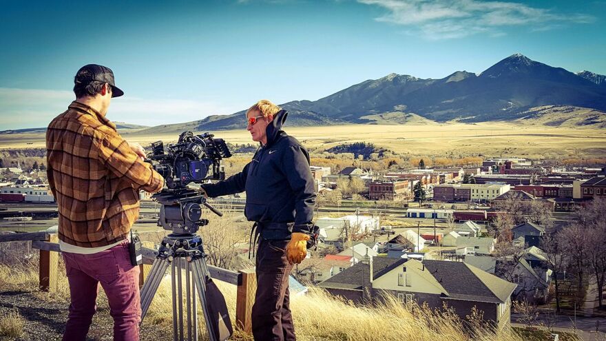 Two people fiddle with a large camera on a beefy tripod. They're on a hill overlooking a town with mountains in the background.