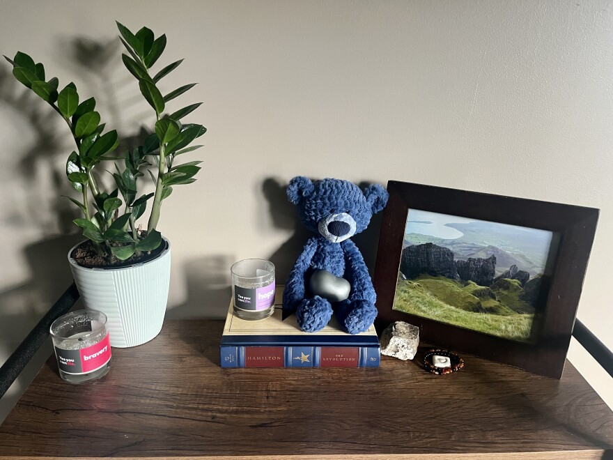 A shelf holds two candles, a picture of a landscape and a blue teddy bear. The bear is holding what looks like a small, gray heart-shaped rock in his lap. 
