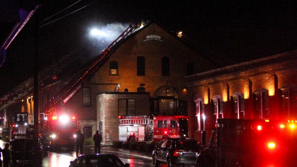 Emergency vehicles, including two fire trucks, are parked outside a large brick building on a dark, wet night. Smoke billows from the pointed roof, while a firefighter works on a tall ladder. Two other firefighters look on from the street below. 