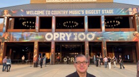 A man standing in front of a large arena with signs: "Grand Ole Opry House", "Welcome to Country Music's Biggest Stage", and "Opry 100."