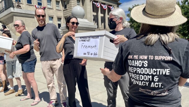 People lined up on the steps of the Idaho Capitol on a sunny day, handing each other white cardboard boxes. One woman with her back to the camera to the forefront right wears a t-shirt with the words "Ask me how to support Idaho's Reproductive Freedom and Privacy Act." She's just handed a box with the word "Fremont" sharpied on it to 