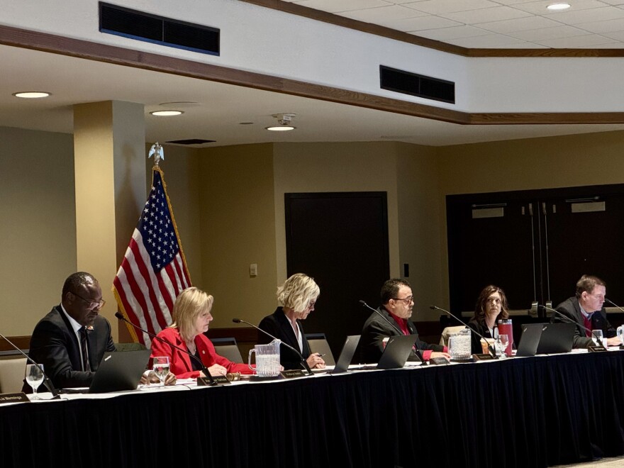 Six people sit at a long table with laptops and papers during a formal meeting. An American flag is in the background, and the room has neutral colors and recessed lighting.