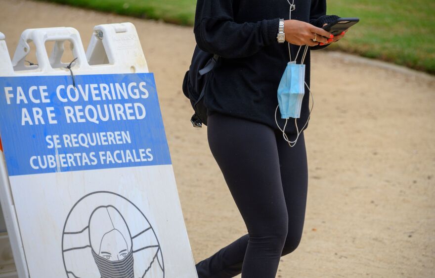 A woman carrying her mask walks past a sign mandating the wearing of face coverings in an effort to control the spread of coronavirus as she walks in Palisades Park in Santa Monica, Calif., on July 25, 2020.