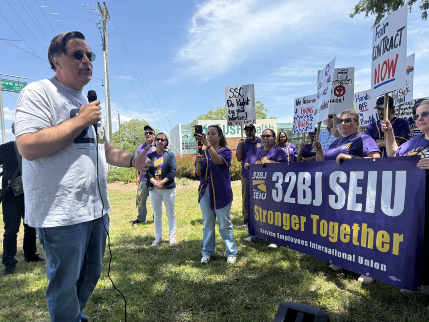 Un hombre con una camisa gris habla por un micrófono ante una multitud de manifestantes con camisas moradas que sostienen carteles.