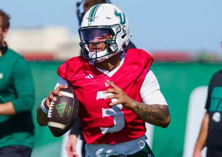 football player in a red practice jersey with number 3 and a white helmet holding a football as he looks to throw