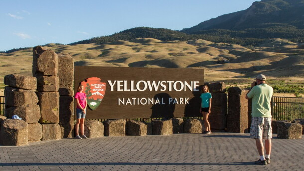 A man takes a picture of two women standing in front of an entrance sign with the words “Yellowstone National Park.”