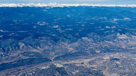 Dry mountains and hills are pictured next to sprawling cities as seen from an aerial photograph. A small ribbon of snow capped mountains are in the background. 