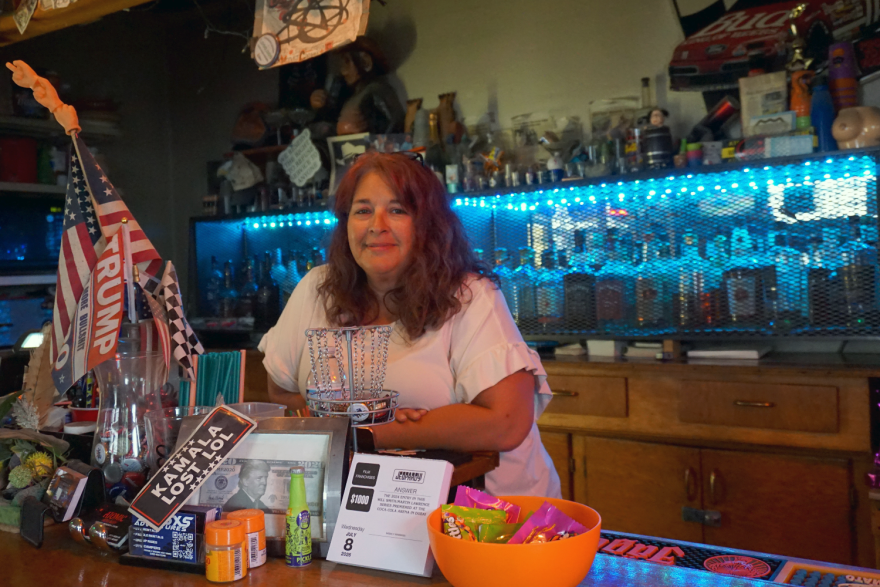 A woman stands behind a bar that's covered in knickknacks and pro-Trump paraphernalia. Behind her are more knickknacks and liquor.