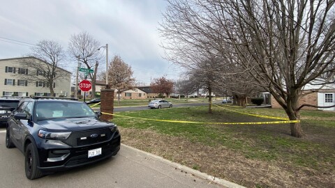 Crime scene tape along several homes and apartment buildings in an urban neighborhood with a police car parked at the curb