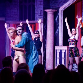 Nine cast members of the Rocky Horror show dance in theatrical clothing on stage during a live performance. 