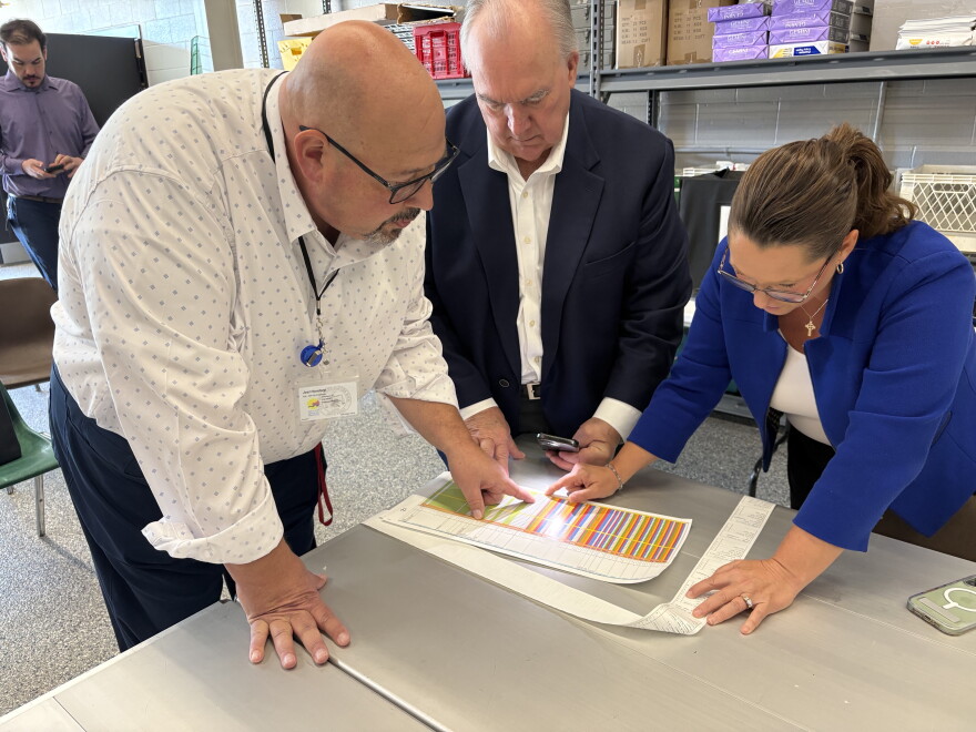 Three people lean over a table, looking closely at a sheet of paper with colored lines.
