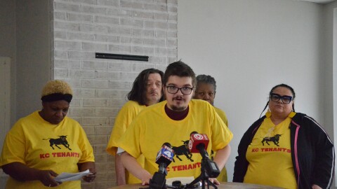 A man in a yellow shirt stands at a table in front of microphones. 