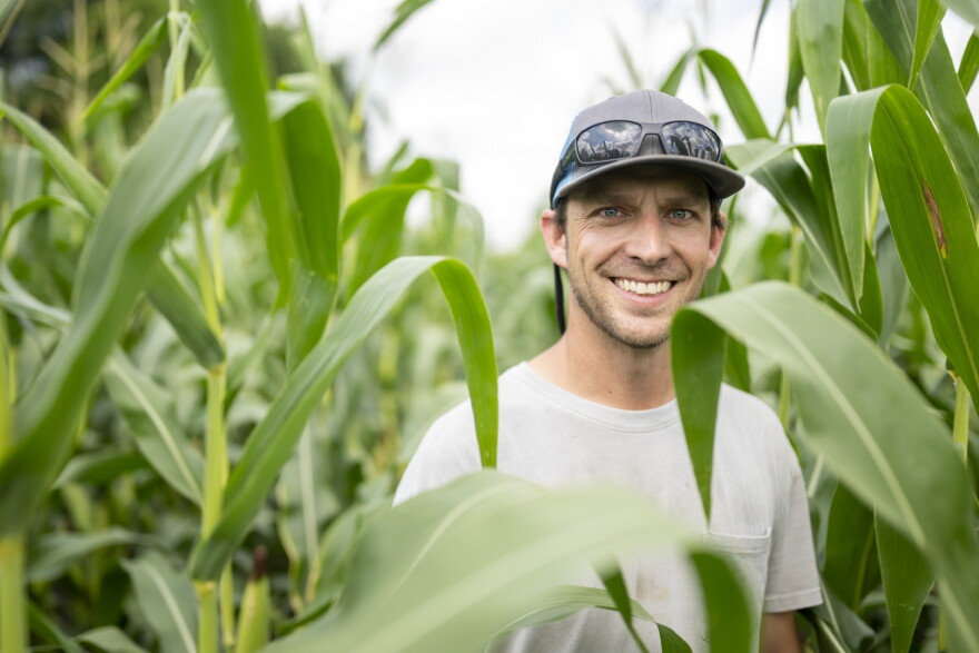 A man wearing a gray baseball hat with sunglasses perched on top and a white t-shirt smiles for a photo in a field of corn.  