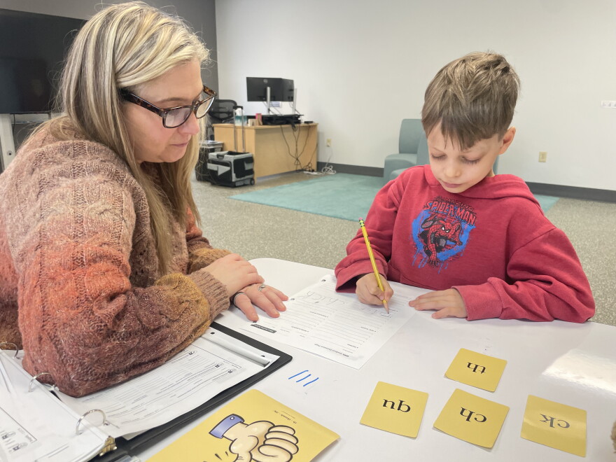 An adult in a sweater and glasses watches a child in a red hoodie complete a worksheet. 