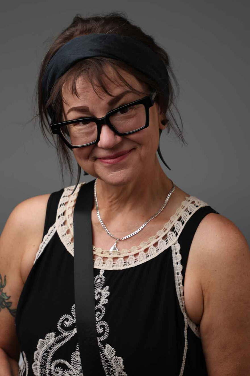 A woman with short brown hair and chunky black glasses smiles at the camera in front of a grey background