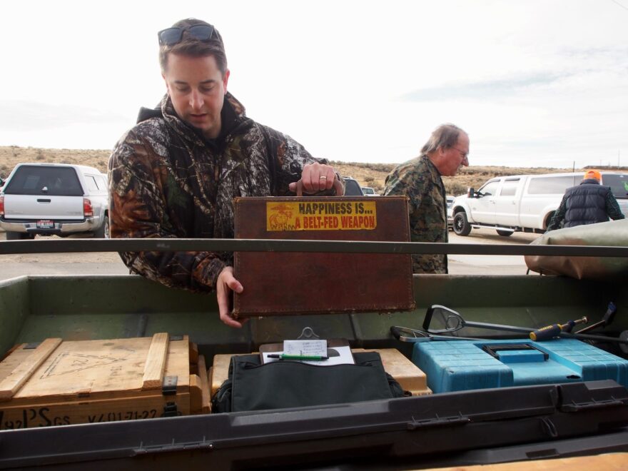 A man wearing a camouflage coat unloads a metal case from the bed of a truck filled with equipment. On the case there's a yello sticker that reads: "Happiness is ... a belt-fed weapon."