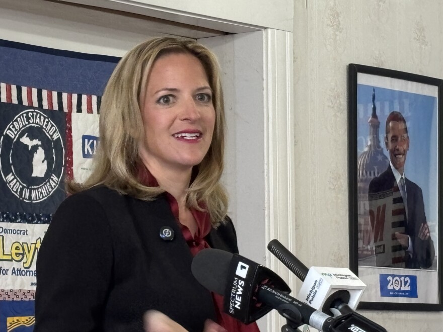 Jocelyn Benson speaks at a press conference, standing indoors near a doorway with microphones in front of her. A campaign poster and a framed photo of former President Barack Obama hang on the wall behind her.