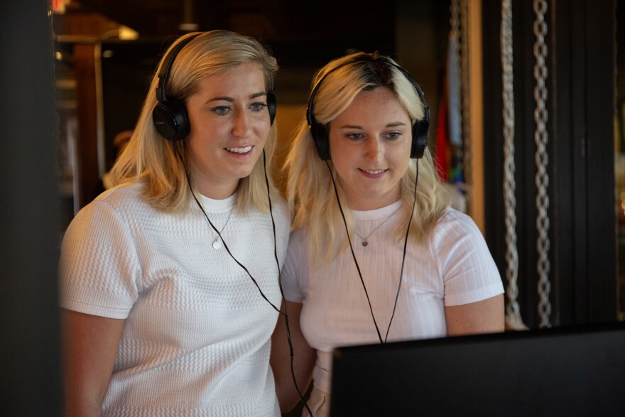 Two blondes in white shirts stand side by side with headphones on looking at a TV display. 