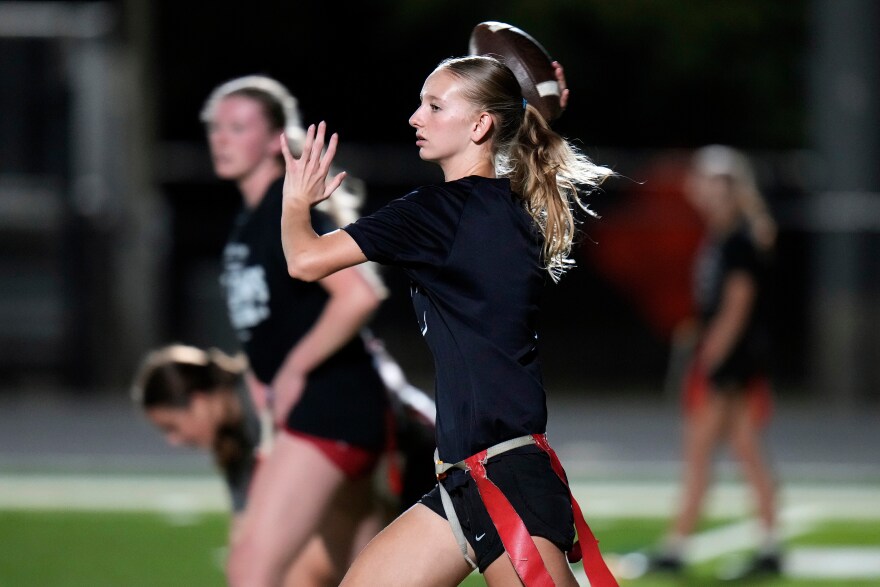 A young girl with a blonde ponytail rears back her right hand to throw a football. It's nighttime. She's wearing a dark uniform shirt and shorts and has a belt with red flags tied around her waist. 