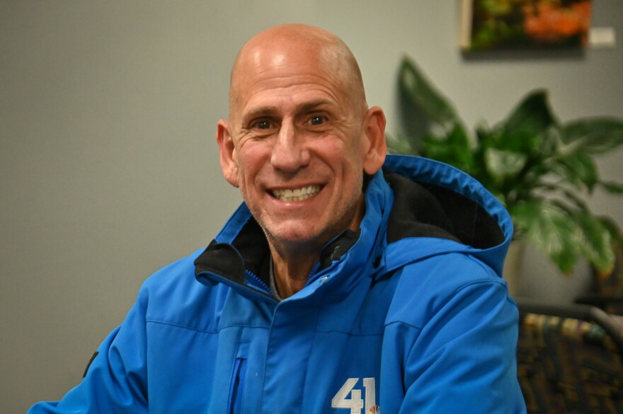 A man wearing a blue coat sits inside an office setting, smiling at the camera.