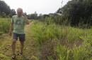 A man in glasses, a light green shirt and gray shorts walks down a path cut through tall green grasses. He's looking down and touching his head. 