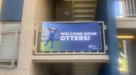 A blue banner with white letters that read "WELCOME HOME OTTERS" on the right of the banner is paired with an image of a college otter mascot standing up with hands raised on the left of the banner. The banner hangs on rails outside a staircase in a college dormitory setting. 
