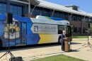 A man stands speaking at a podium, with a large bus behind him. The Connect Transit bus has a hawk on it and says Heartland Community College on the windows.