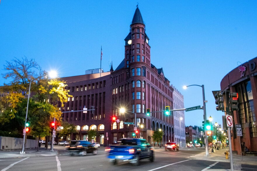 A brick building with a pointed spire is lit at twilight with evening traffic whizzing by.