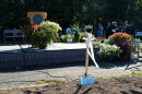 A silver and wood shovel with a white bow is stuck in the brown dirt in front of rises with color flowers around it. 