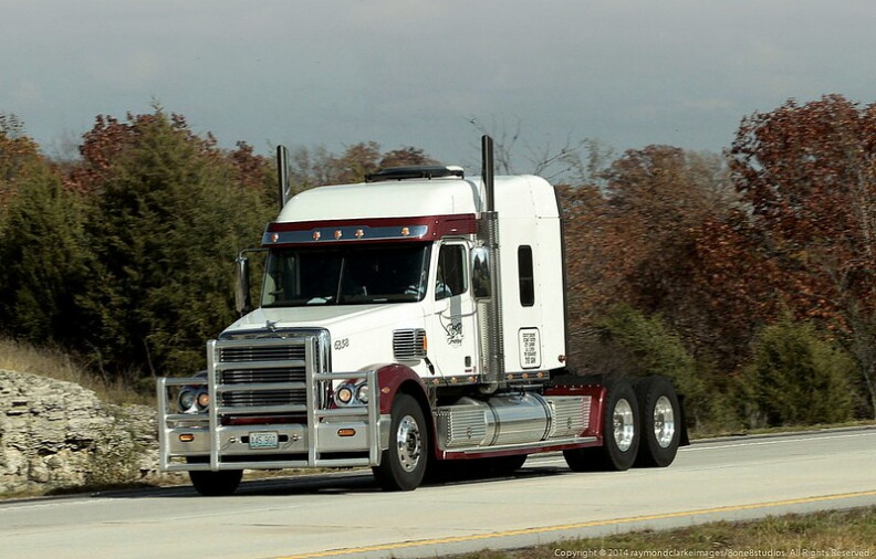 A white semi-truck cab sits on a road in front of greet and dark red trees.