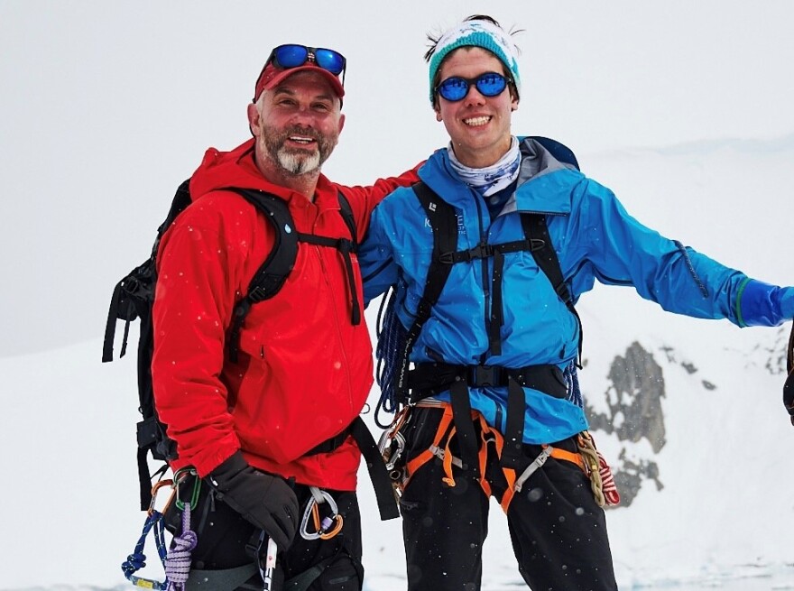 Rob DeLena is in a red jacket and harnesses, with his sunglasses perched on his red hat. He has his arm around his son, Ryan DeLena, who's wearing a blue jacket, a blue and white hat, and sunglasses. They're posing in front of a snowy background.jpg