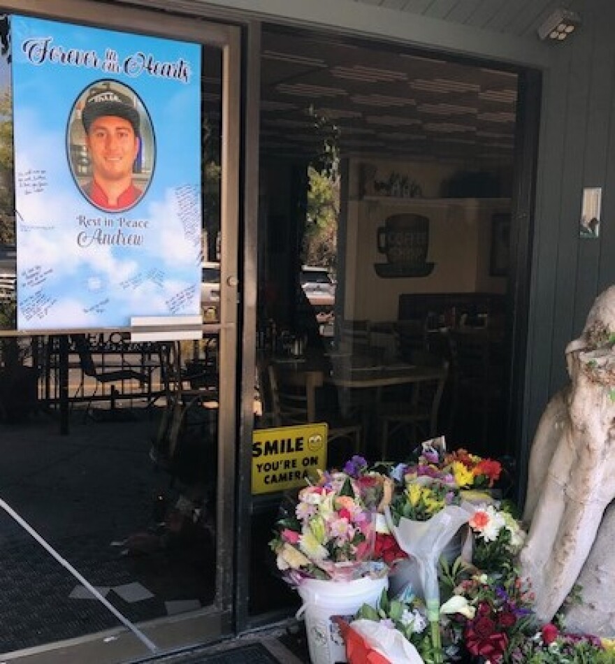 A picture of Andrew Canepa is on a poster on a glass door  of his Newbury Park cafe. There are several bouquets of flowers in front of the window.