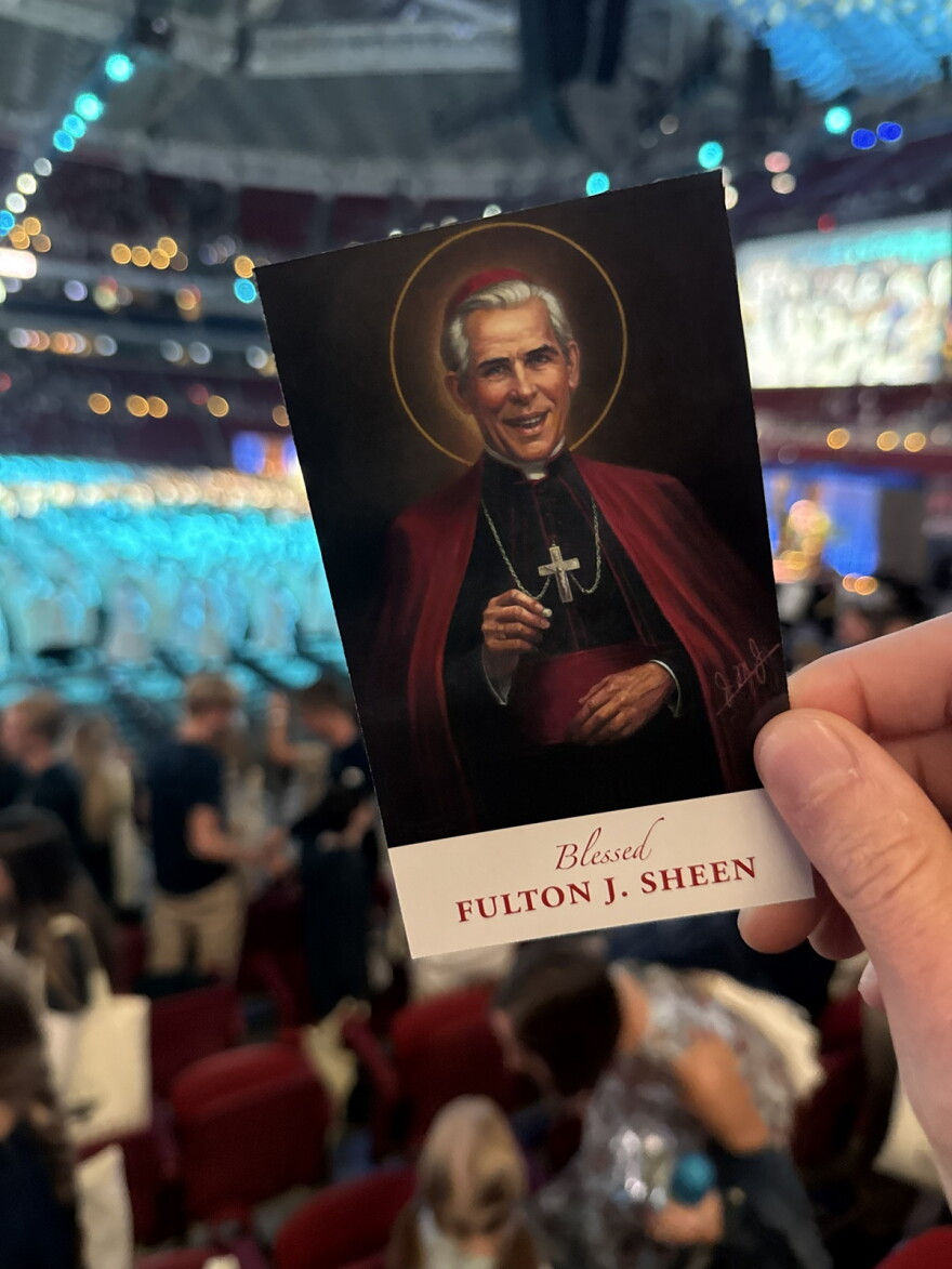 A spectator holds of a card with a portrait of the late Archbishop Fulton Sheen in front of a crowded arena.
