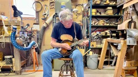 A man in a black t-shirt and jeans sits on a wooden stool in the middle of a workshop to play the mandolin.