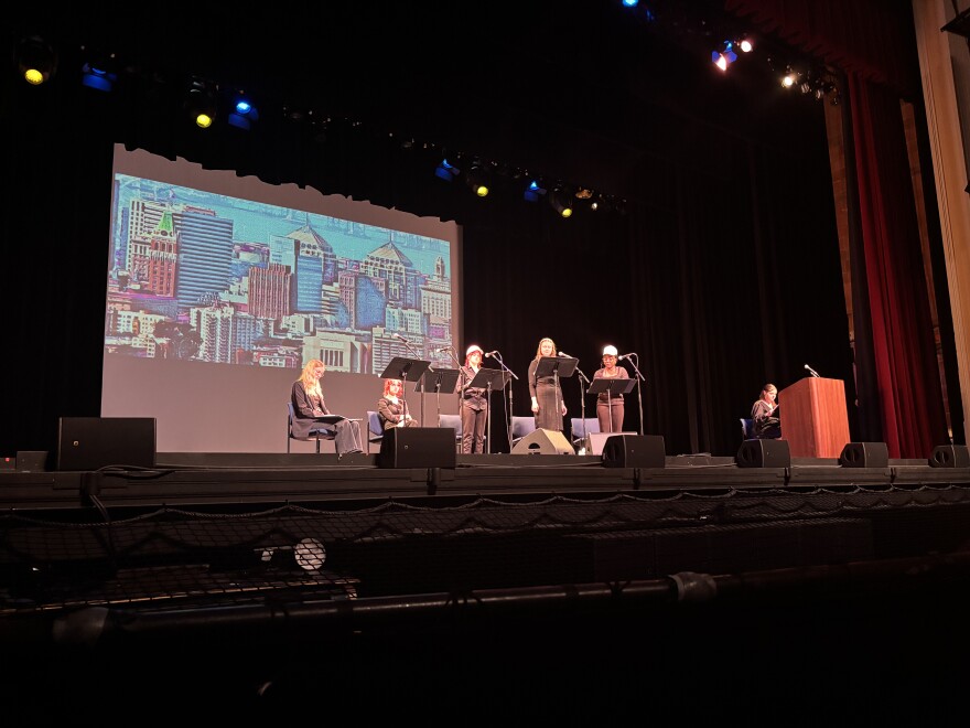 A group of students read their lines from scripts on a dark stage with a building skyline image projected behind them.