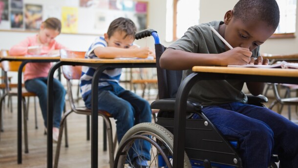A stock image of children writing on desks in a classroom. An African-American student in a wheelchair sitting closest to the camera is very focused on his work. There are two children behind him in the background also writing.