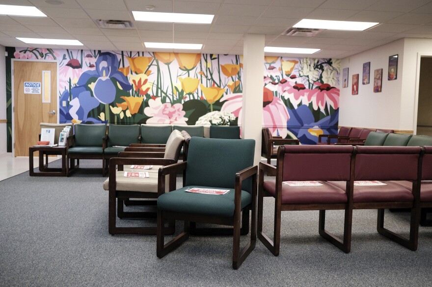 Empty green and maroon chairs sit in a waiting room with a colorful flower mural on one wall.