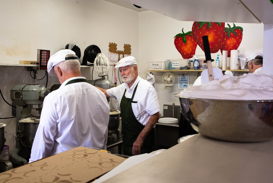 Men in hats and aprons stand in a kitchen preparing baked goods. 