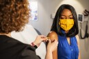 A woman with curly hair places a Band-Aid on the arm of another woman wearing a yellow mask.