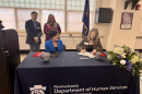 Kimberly Tenbus, CEO of Clarks Summit State Hospital (right), signs the clinical agreement while Pa. Human Services Sec. Val Arkoosh (left, seated), Dr. Ryan Smith, the associate dean of clinical affairs and graduate medical education at IUPCOM (standing left) and IUPCOM's founding dean Dr. Miko Rose watch.
