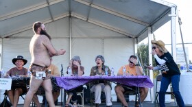 A hairy man walks past a table of people laughing. 