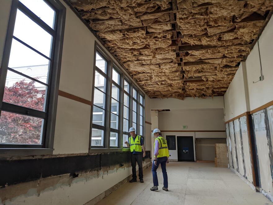 Two men in safety vests and hard-hats look out a window on the second floor of an old school. The school is under renovations, and the ceiling is exposed, showing the insulation.