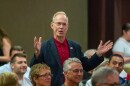 Man standing in a crowd in a meeting room while wearing a dark sports jacket and red collared shirt and extending his hands as he speaks. 