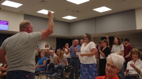 A man in a grey shirt faces a crowd of clapping and standing people. He has his fist in the air as a sign of victory and other people pump their fists as well. The people are in a school cafeteria. 