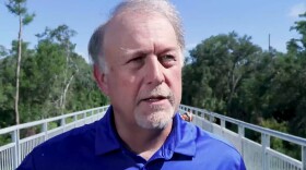 Man with grey hair and blue polo stands outside with bridge behind him 
