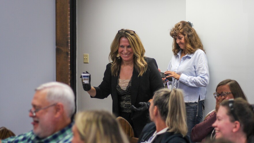 A well-dressed woman in a courtroom smiles as she holds a closed cup of urine in gloved hands. 