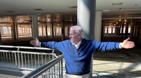  Ken Greene, director of commercial development for Sibley Square, gestures with arms wide, explaining how the fifth-floor of Sibley Square will be divided up for the future offices of Bandwidth, other common areas and still-unleased space. The sun is shining in through a large, glass atrium room. He is standing alongside the railing. 